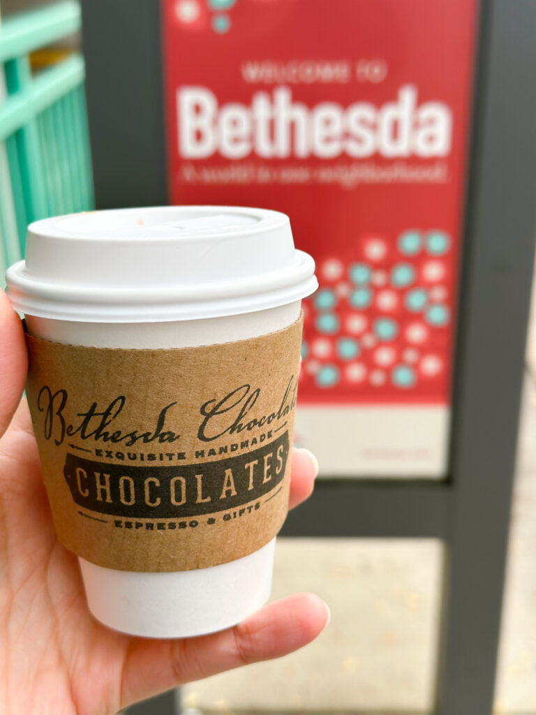 A cup of hot cocoa from Bethesda Chocolates held up in front of a “Welcome to Bethesda” neighborhood sign.