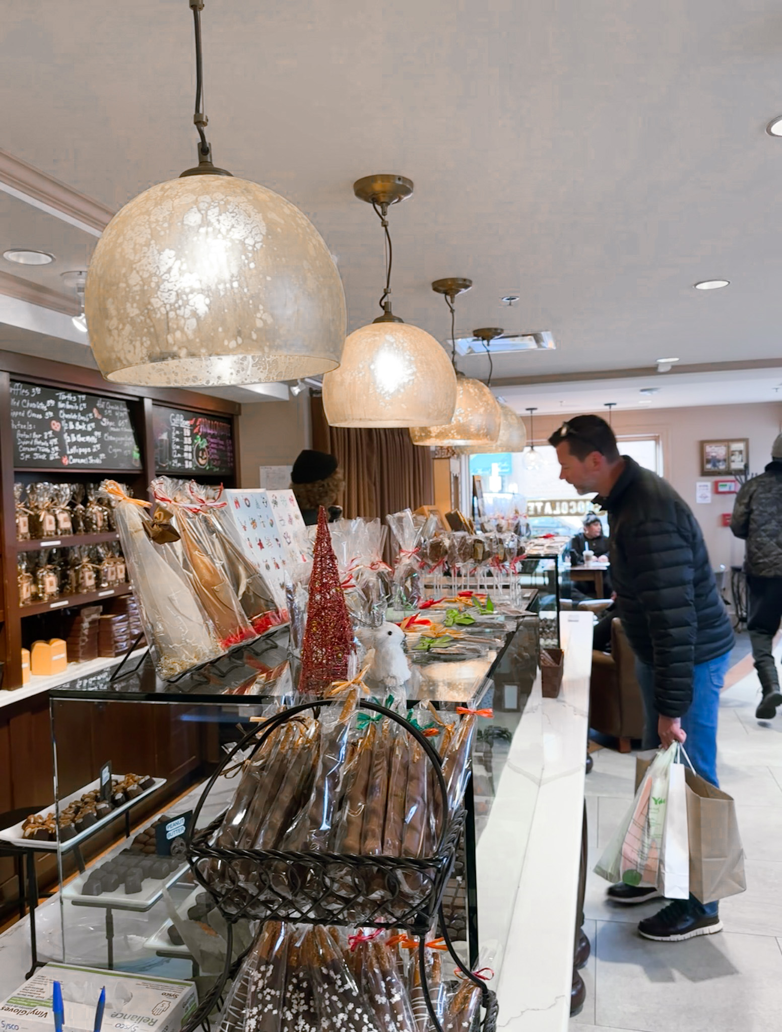 Interior of Bethesda Chocolates with dome pendant lights, customers, and a glass counter filled with wrapped chocolate treats.