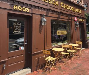 Exterior of Bethesda Chocolates on Woodmont Avenue in downtown Bethesda with yellow cafe tables and chairs.