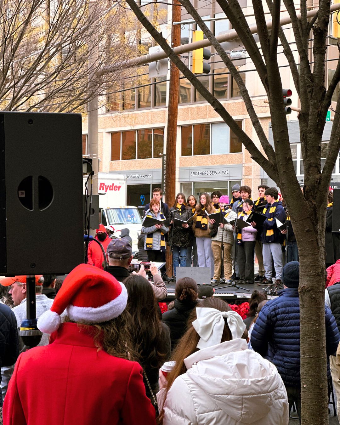 Two young women in the audience crowd wearing a Santa hat and a large bow, watch the choir performance.