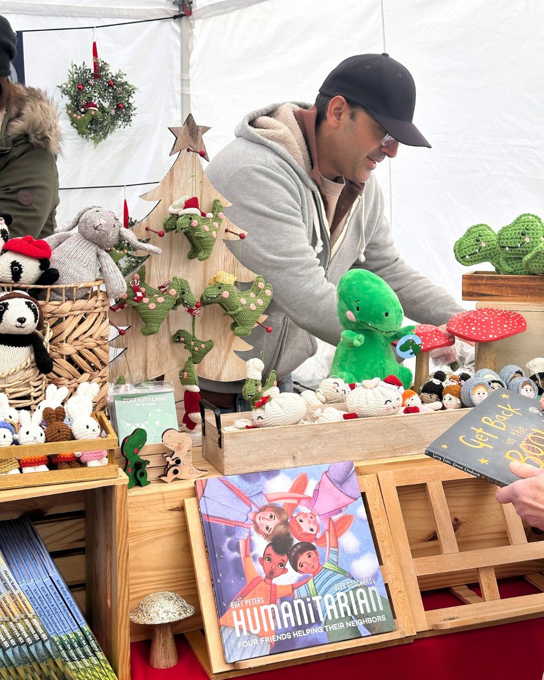 Vendor arranges crocheted dinosaurs and toys, behind display of childrens books.
