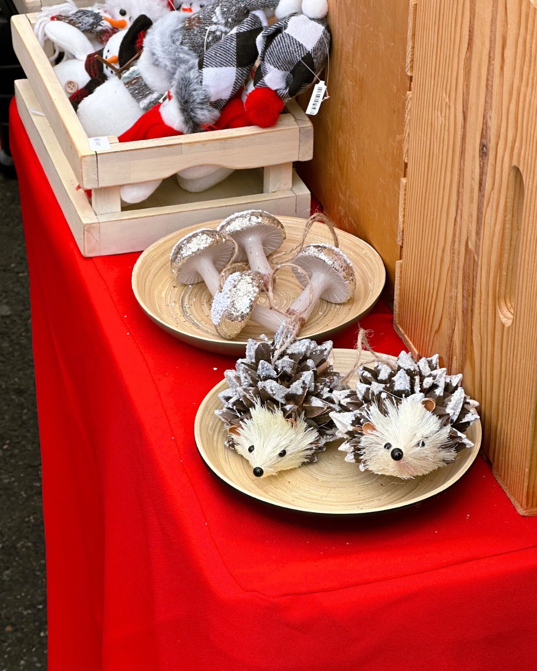 Pinecone hedgehog ornaments, glitter mushrooms, and winter themed stuffed animals displayed on red tablecloth.