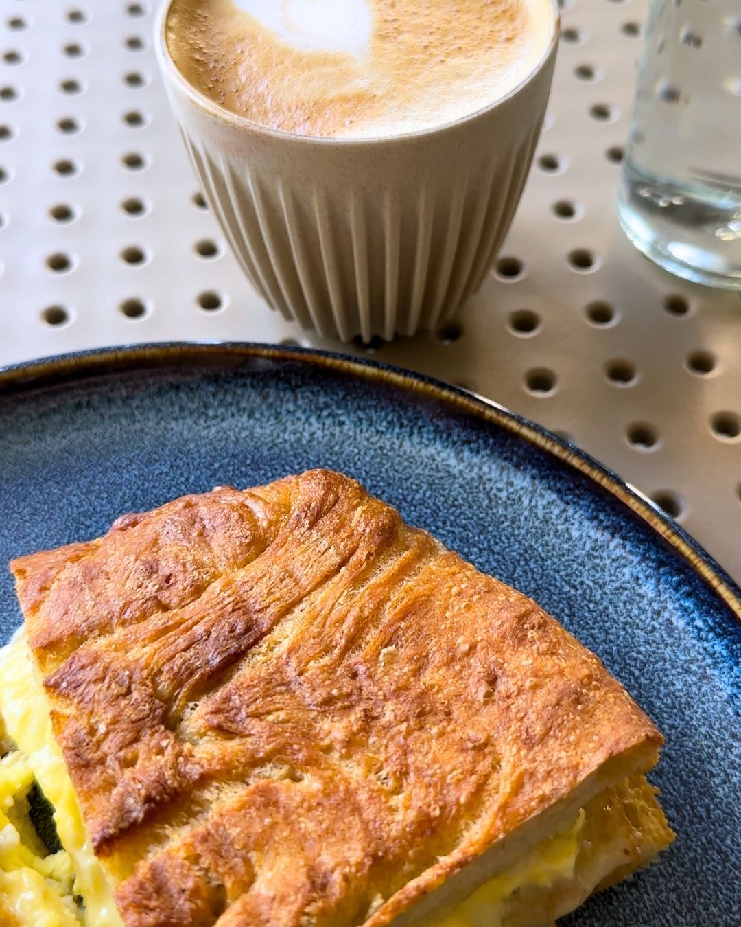 The Wilson breakfast sandwich on house-made focaccia with scrambled eggs and cheddar on a textured ceramic plate next to a latte