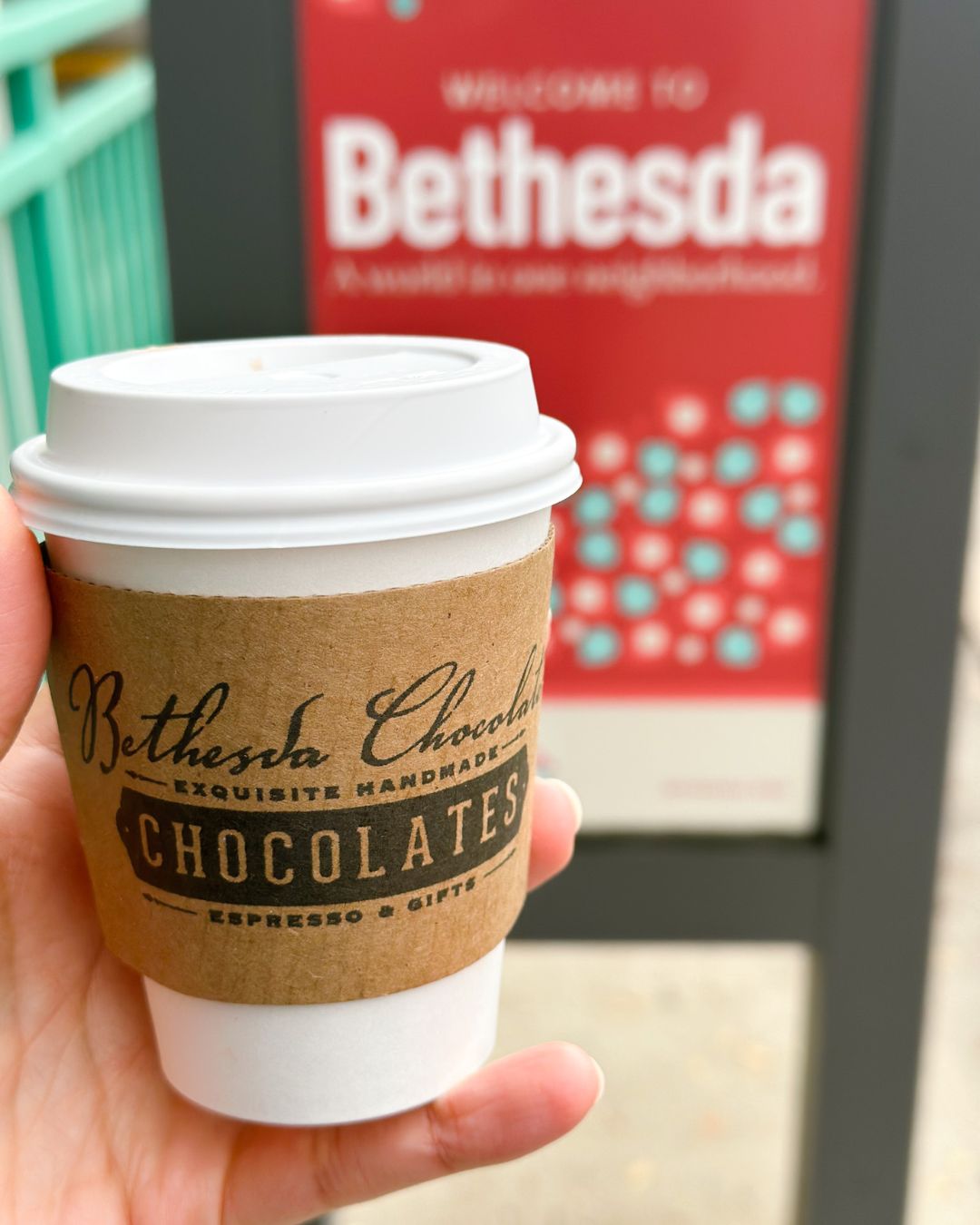 A cup of hot cocoa from Bethesda Chocolates held up in front of a Welcome to Bethesda sign.