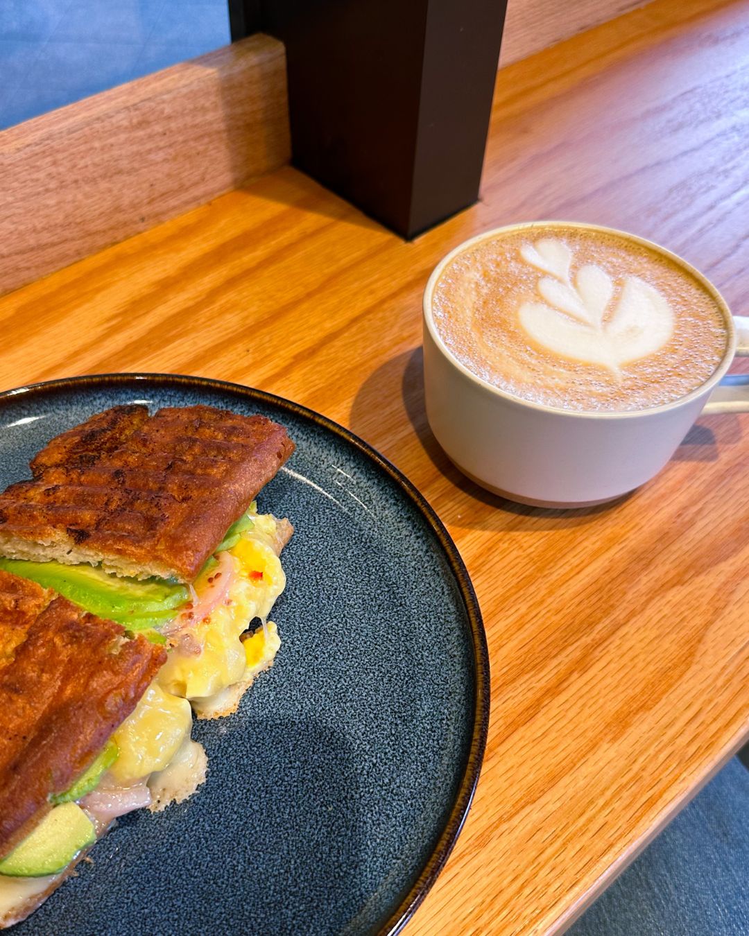 A cup of coffee with latte art next to the Franklin breakfast sandwich on house-made focaccia with: avocado, scrambled eggs, cheese, and pickled onions.