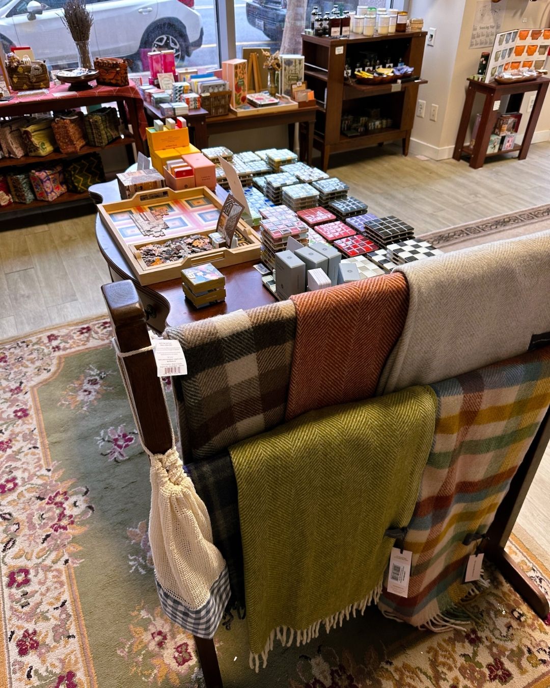 Interior of Green & Bean Boutique with puzzles, card games, and colorful ceramic tile coasters on a coffee table, next to thick wool scarves hanging nearby