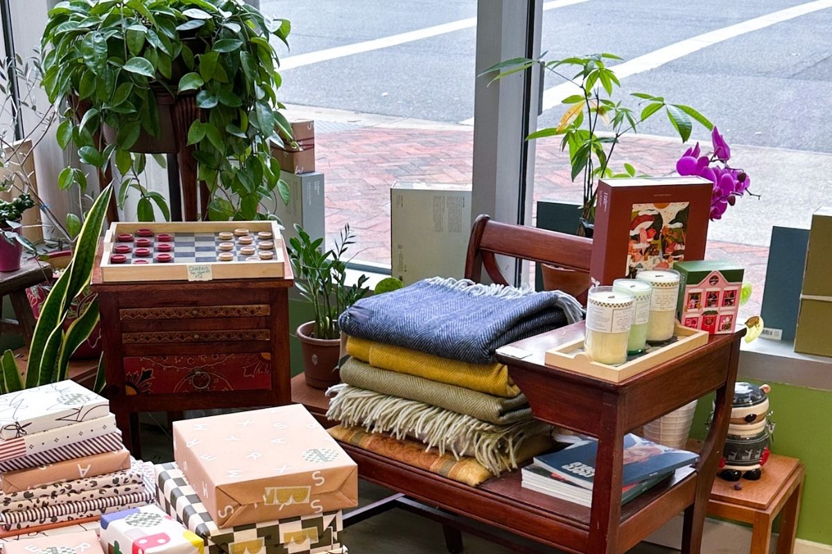 Thick wool throw blankets on a small vintage wooden bench, decorative wrapped gifts on a coffee table, a checkers set and potted plants by the window