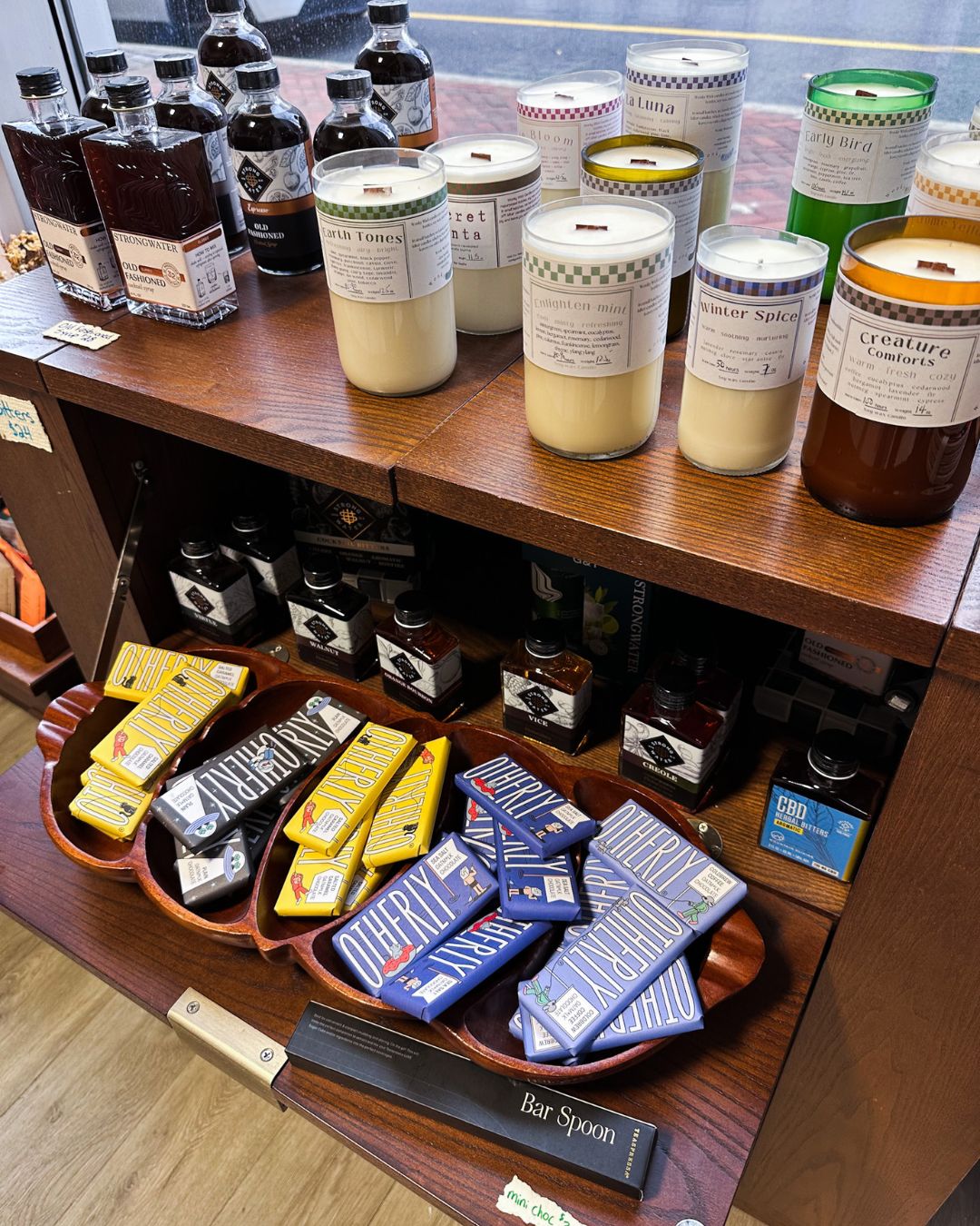 Candles with wooden wicks, flavored syrups and bitters, and Otherly chocolate bars displayed on wooden shelves