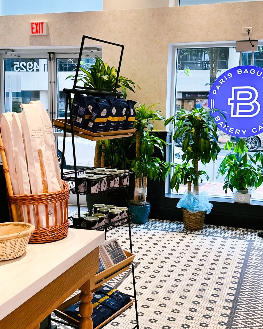 Paris Baguette interior with branded signage on the window, a basket of baguettes, a rack with bags of coffee beans and jars of jam, plants and patterned tile floor
