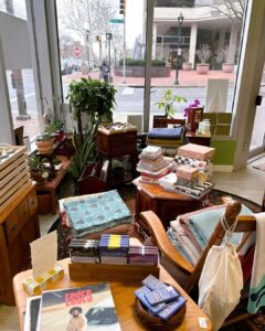 View inside Green & Bean Boutique showing potted plants, folded wool throw blankets, and home goods displayed on vintage wooden furniture near the window